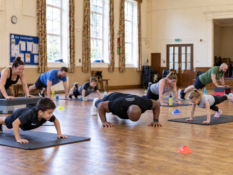 Group fitness class with multiple adults performing plank exercises on yoga mats in a bright studio with tall windows.